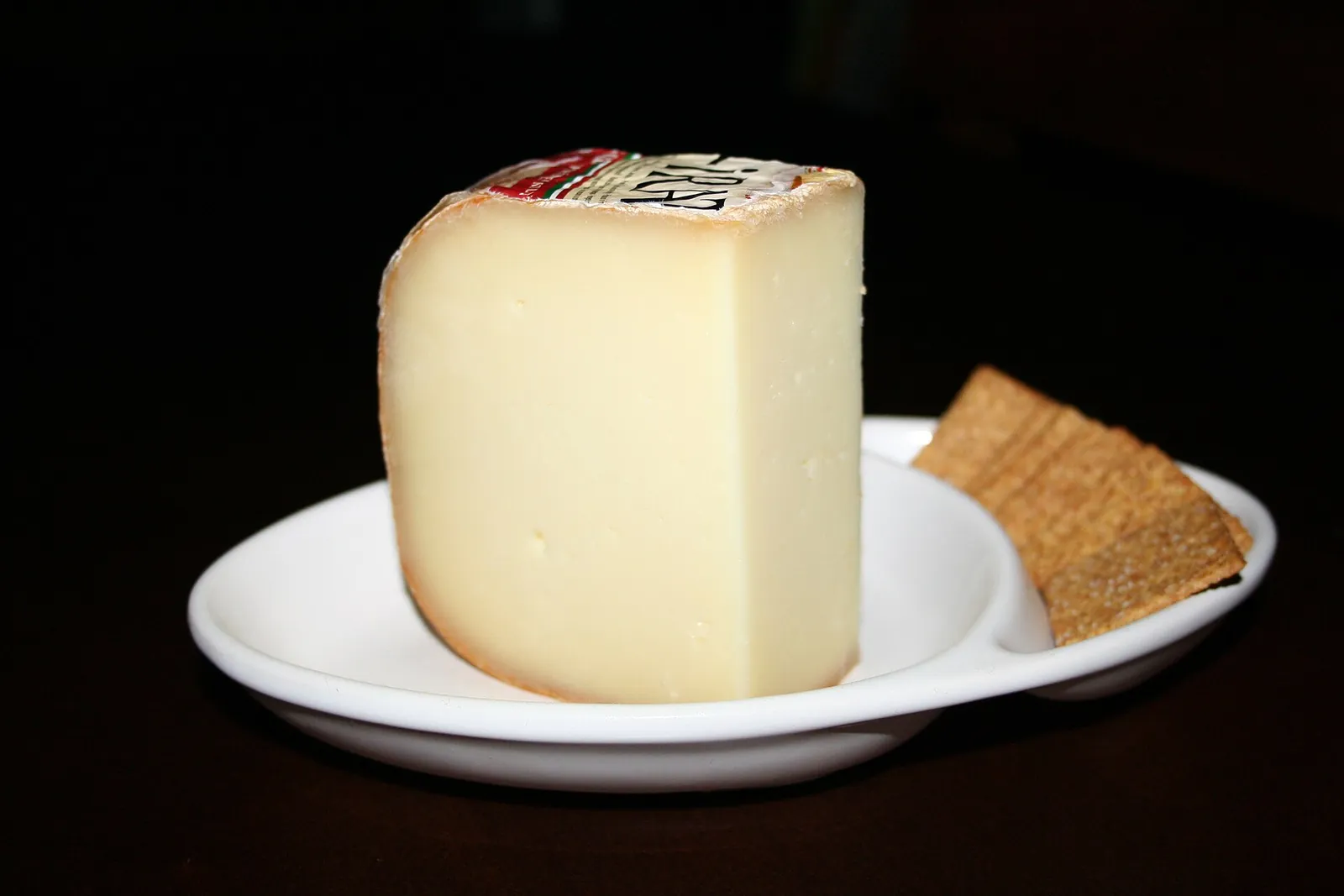 A wedge of Ossau-Iraty sheep milk cheese on a white dish with a cracker, against a black background