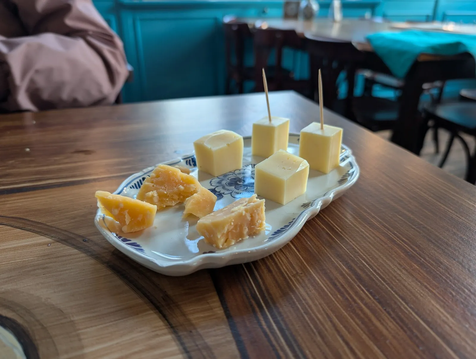 Pale cheese cubes on toothpicks beside amber chunks of aged cheese on a small blue and white dish on a wooden table.