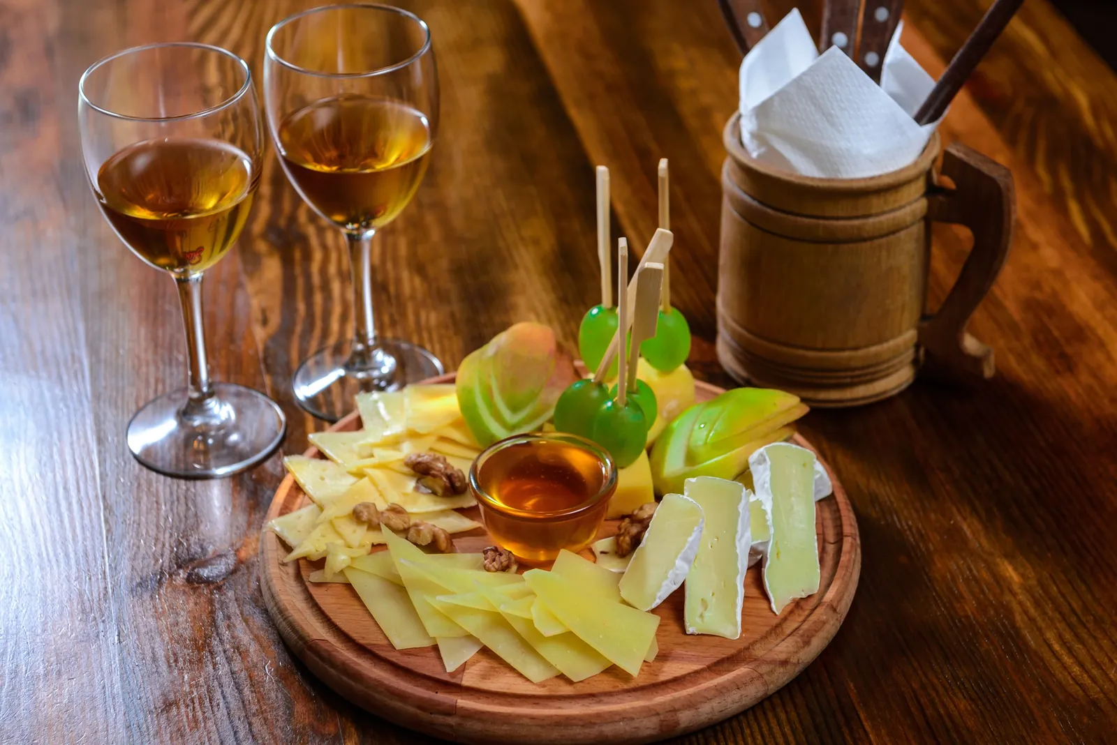 A round board of thin firm cheese slices, brie batons, grapes on skewers, apple slices, walnuts and honey beside two glasses of white wine.