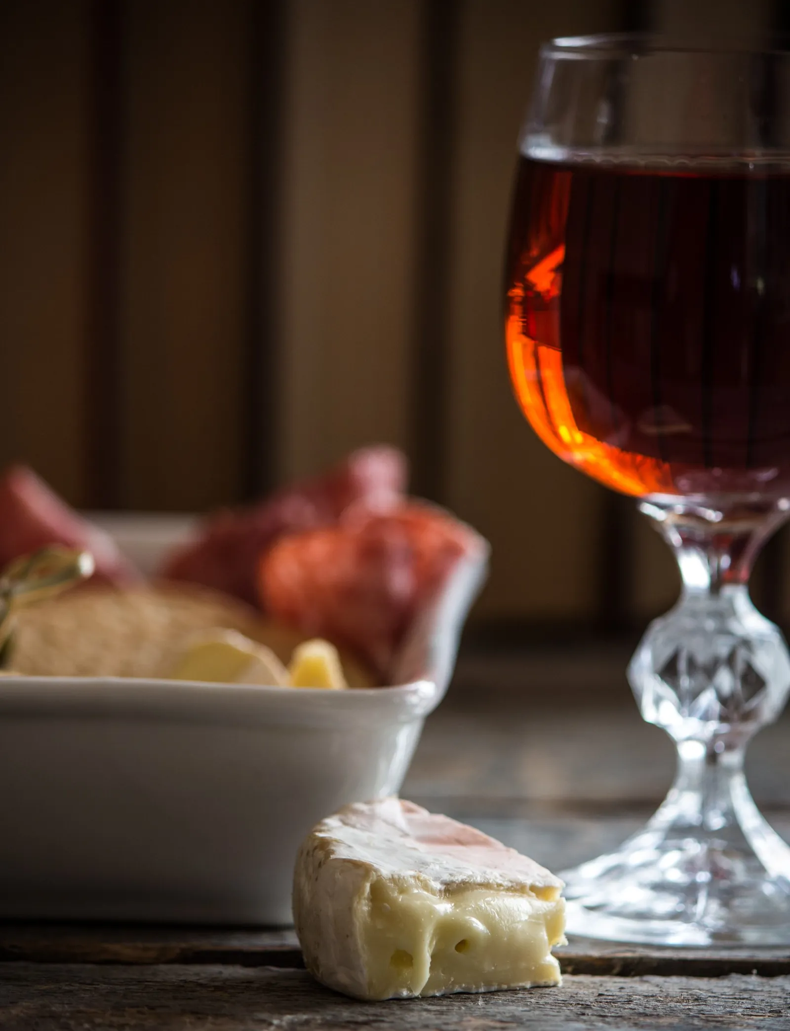 A wedge of soft bloomy-rind cheese in front of a stemmed glass of amber wine, with a dish of sliced salami and cheese blurred behind.