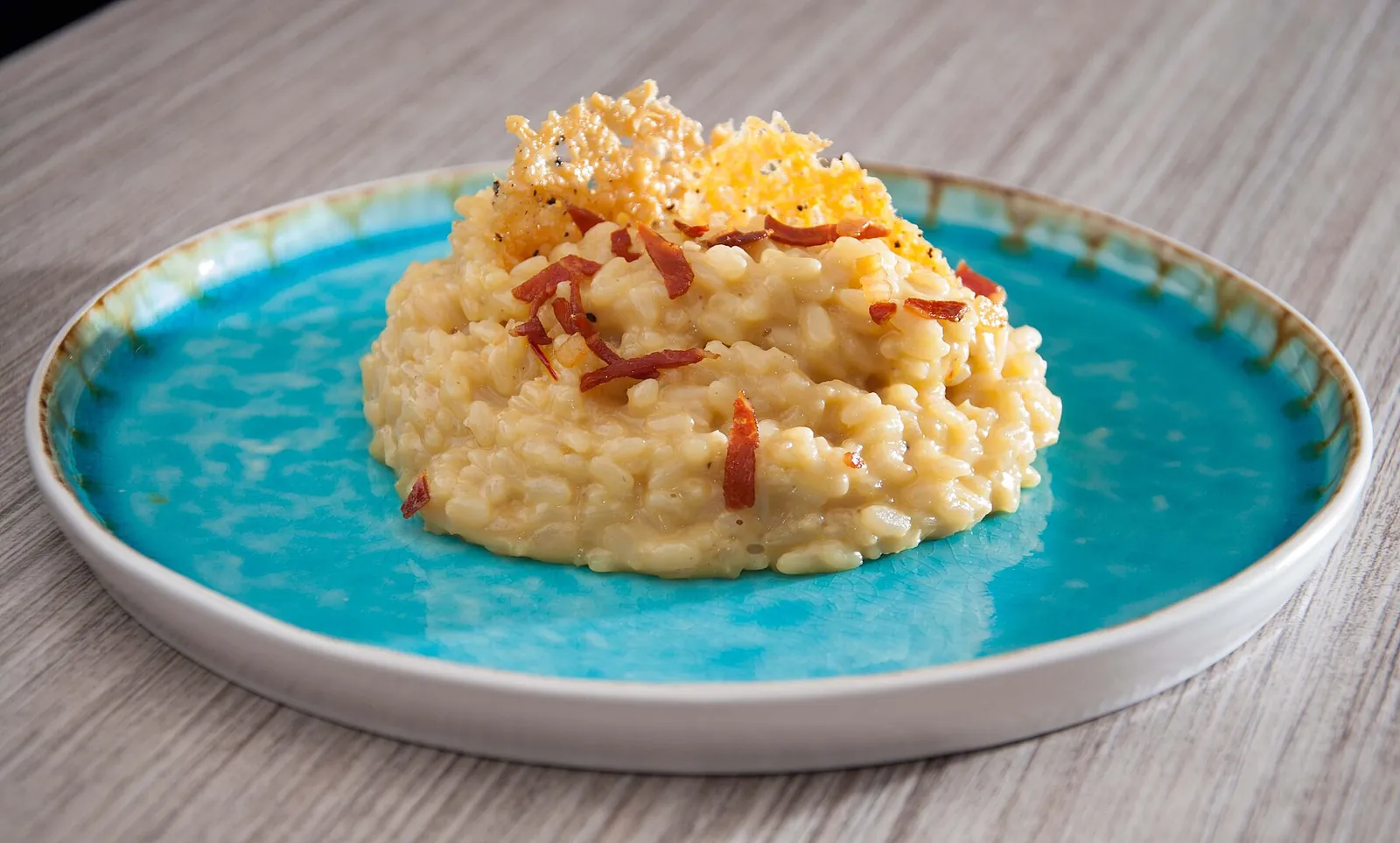 A mound of creamy risotto on a turquoise plate, topped with a crisp golden Parmesan wafer and small crisp pieces of bacon.