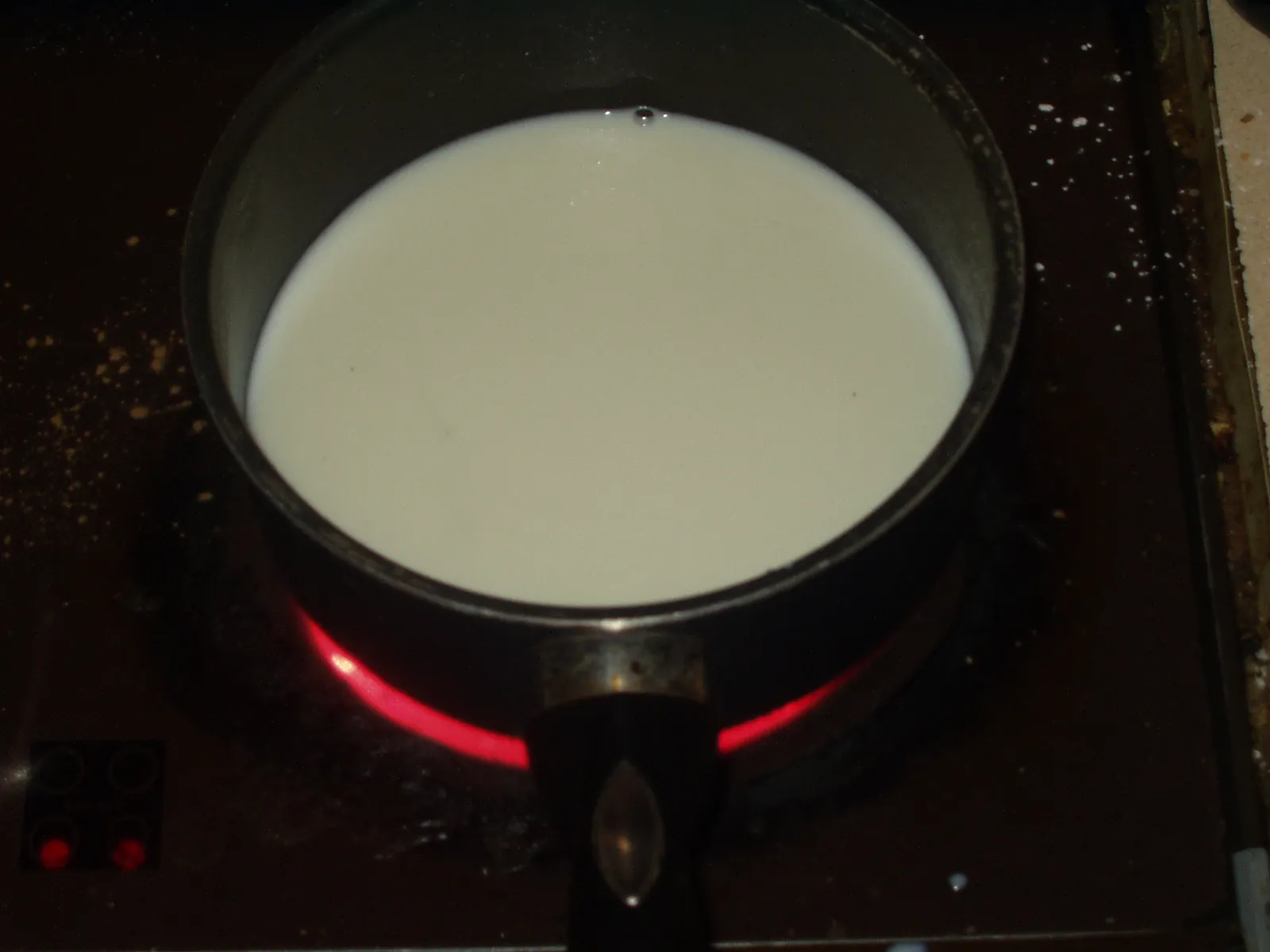 Fresh white milk in a dark saucepan on a stovetop, with the red glow of the heating element below the pan.