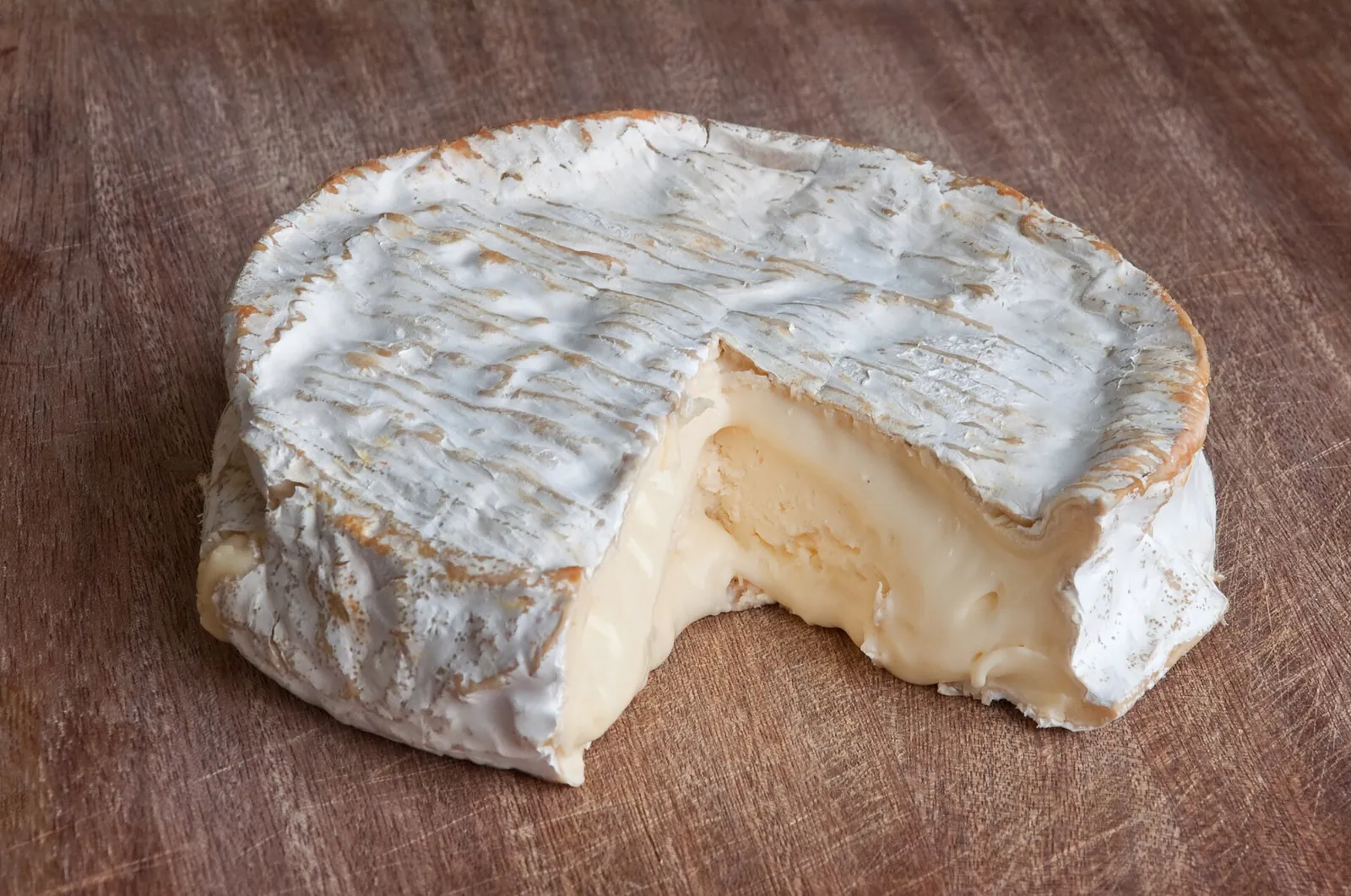 Raw milk Coulommiers cheese with a wedge cut away on a wooden table, showing the white bloomy rind and soft creamy paste.