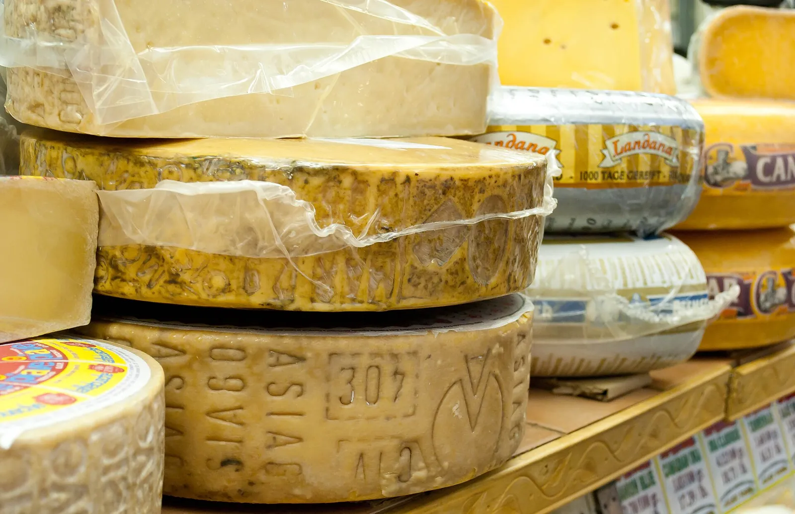 Aged wheels of Asiago stacked on a market shelf, the rinds embossed with the cheese name and a number, with wrapped wheels behind them.