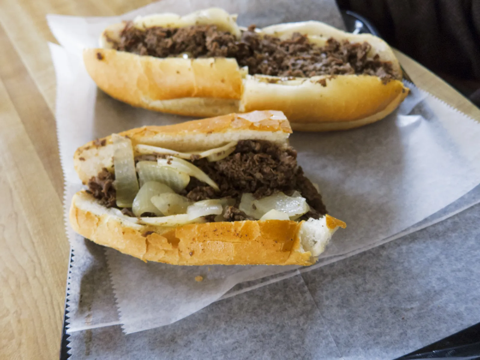 Two halves of a Philly cheesesteak on a hoagie roll, packed with chopped steak, grilled onions and melted cheese, on paper