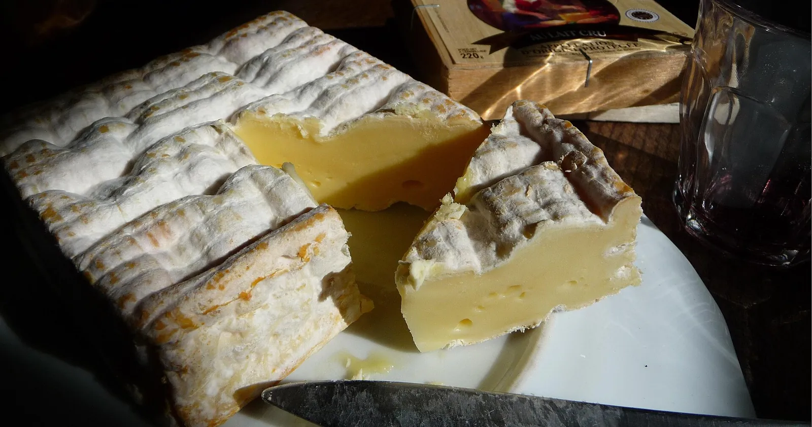 A square Pont-l'Eveque cut open to show creamy yellow paste and a white and orange rind, beside a knife
