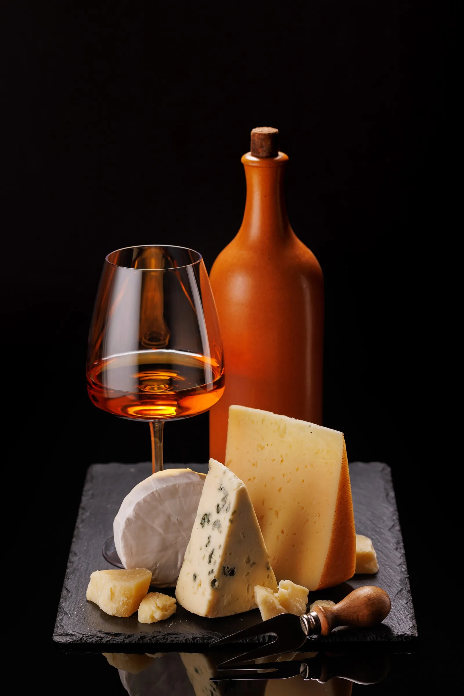 A glass of amber dessert wine and a clay bottle behind a wedge of blue cheese, a small bloomy round and a firm wedge on black slate.