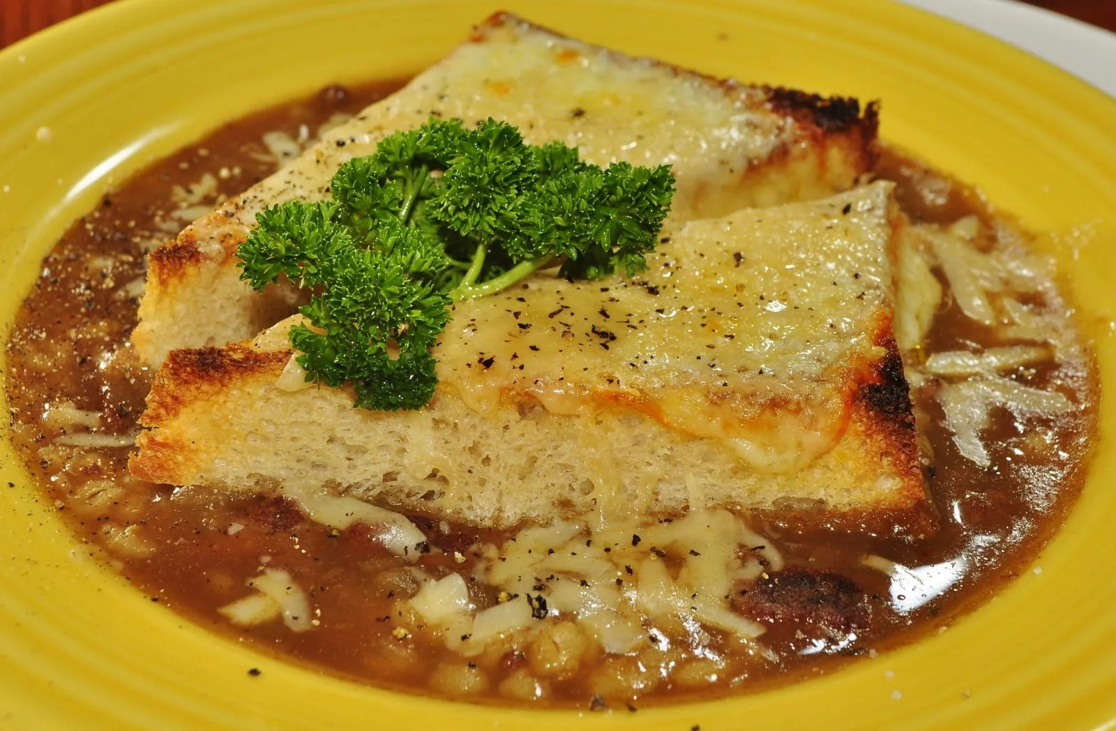 A wedge of toasted bread on a bowl of soup, topped with browned, bubbly melted cheese and a sprig of parsley.