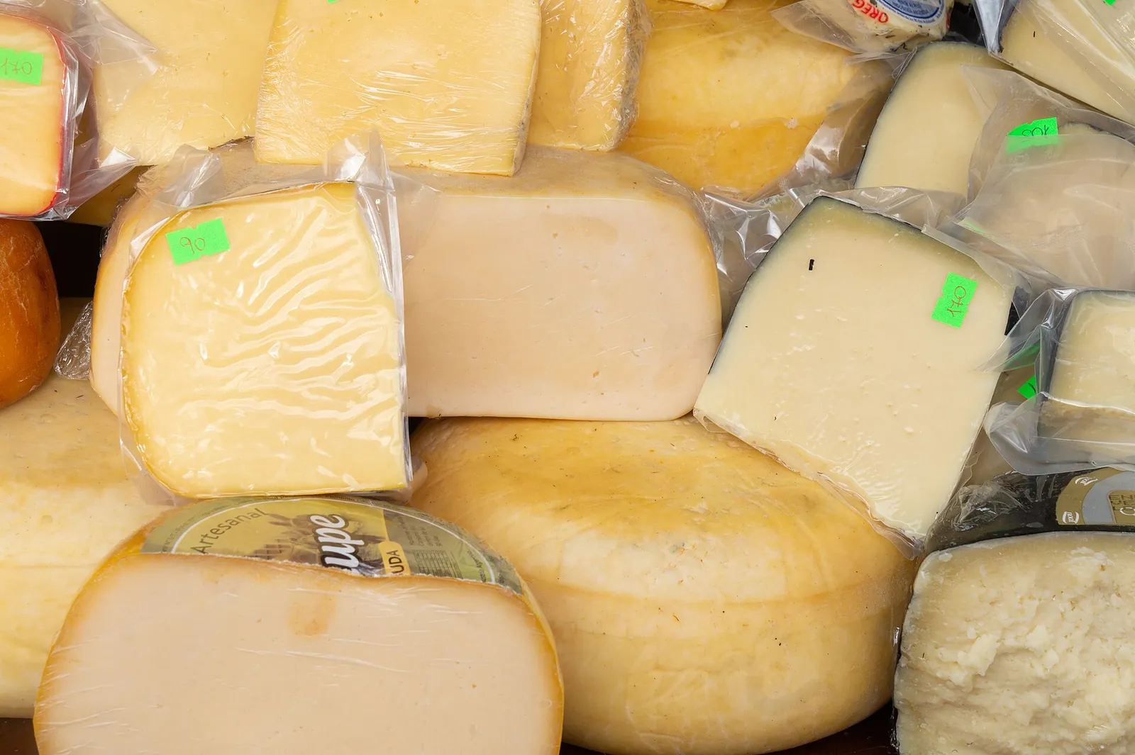 Wedges and wheels of cheese piled together in a market display, many sealed in plastic wrap with small green price stickers