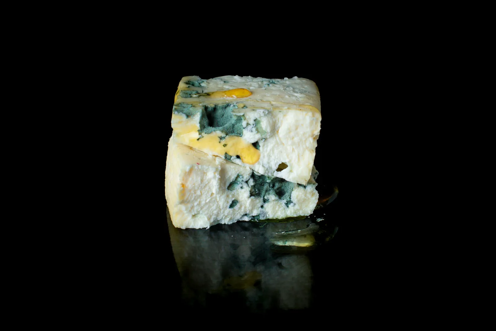 A wedge of blue cheese spotted with fuzzy green surface mold and yellowed patches, sitting on a black reflective surface.