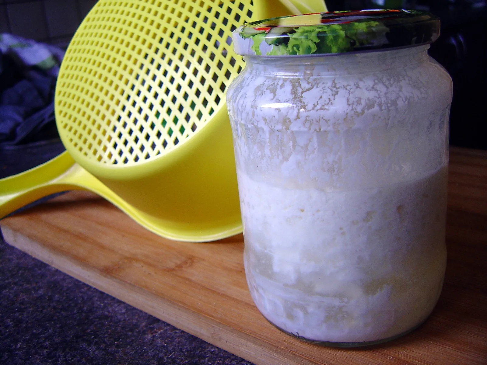 A large glass jar of fermented milk with thick white cultures, set on a wooden board beside a yellow plastic colander.