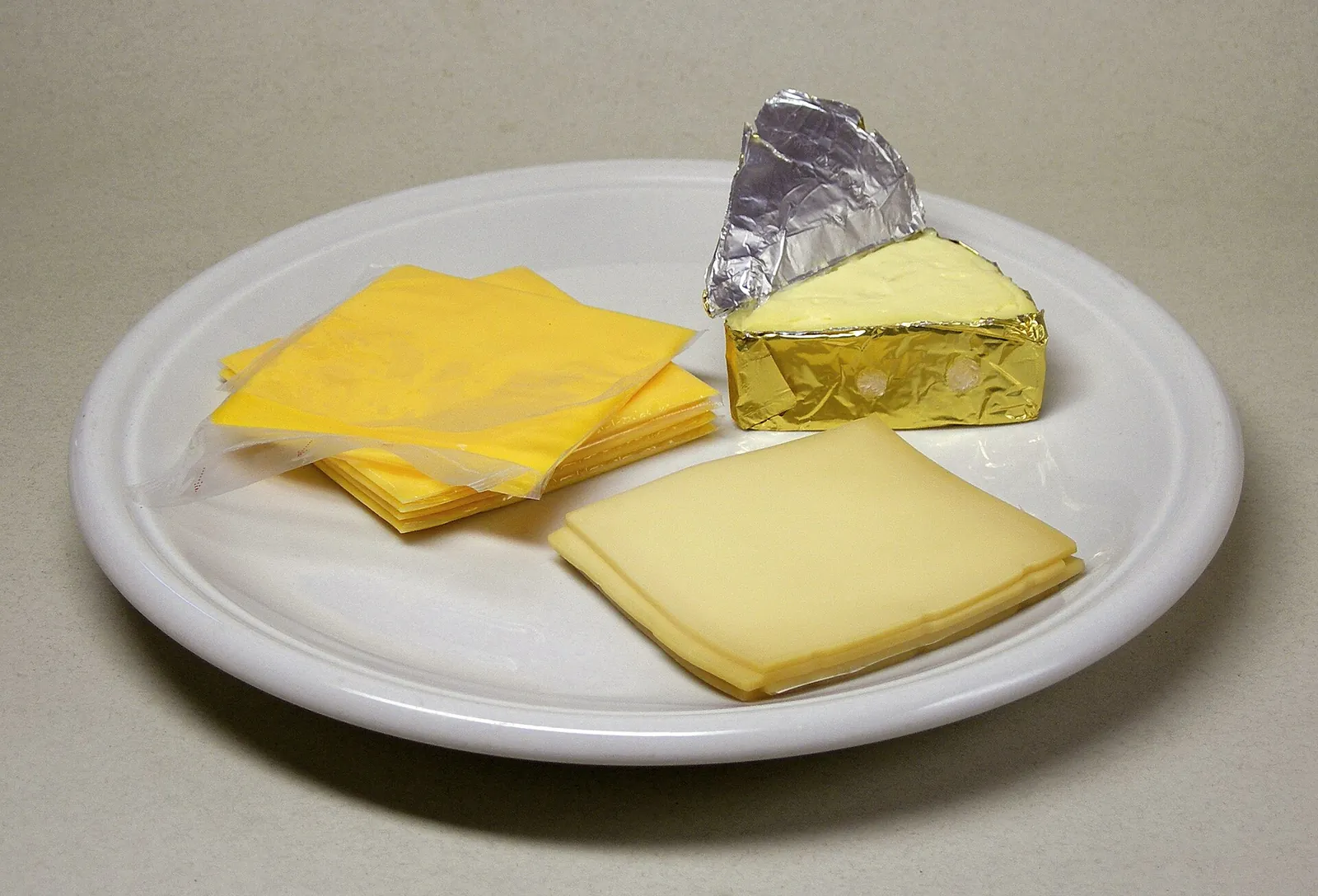 Plate holding a stack of bright yellow processed cheese slices beside a foil wrapped wedge of natural cheese and a pale natural cheese slice.
