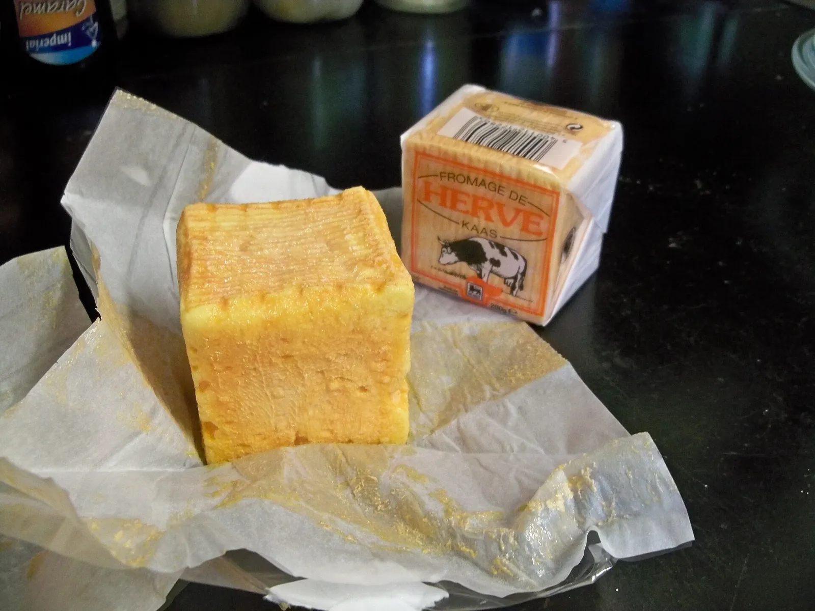 A cube of Herve cheese with an orange washed rind, unwrapped on paper next to a boxed piece of the same cheese