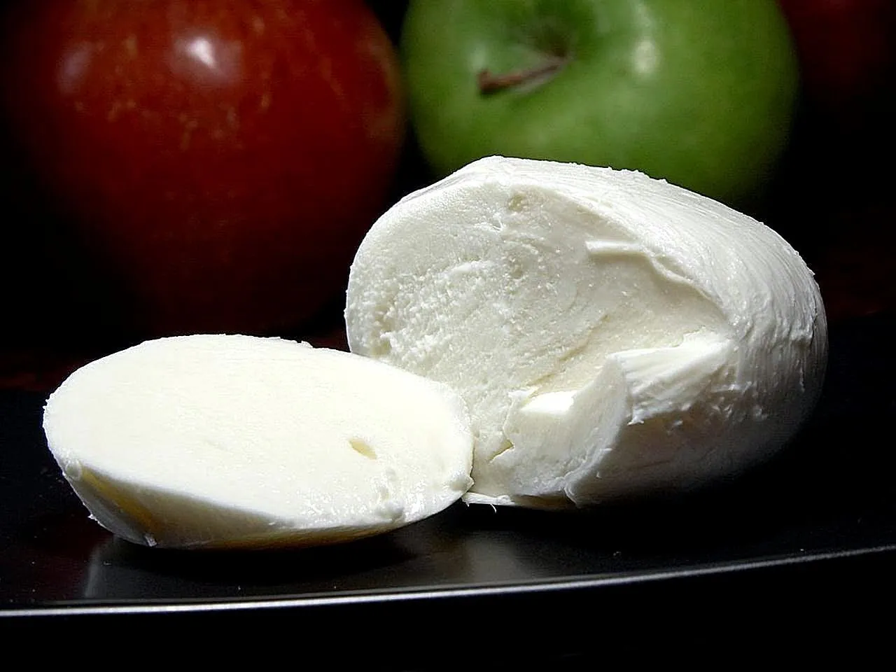 A fresh white mozzarella ball cut open to show the smooth, moist interior, beside green and red apples on a dark surface