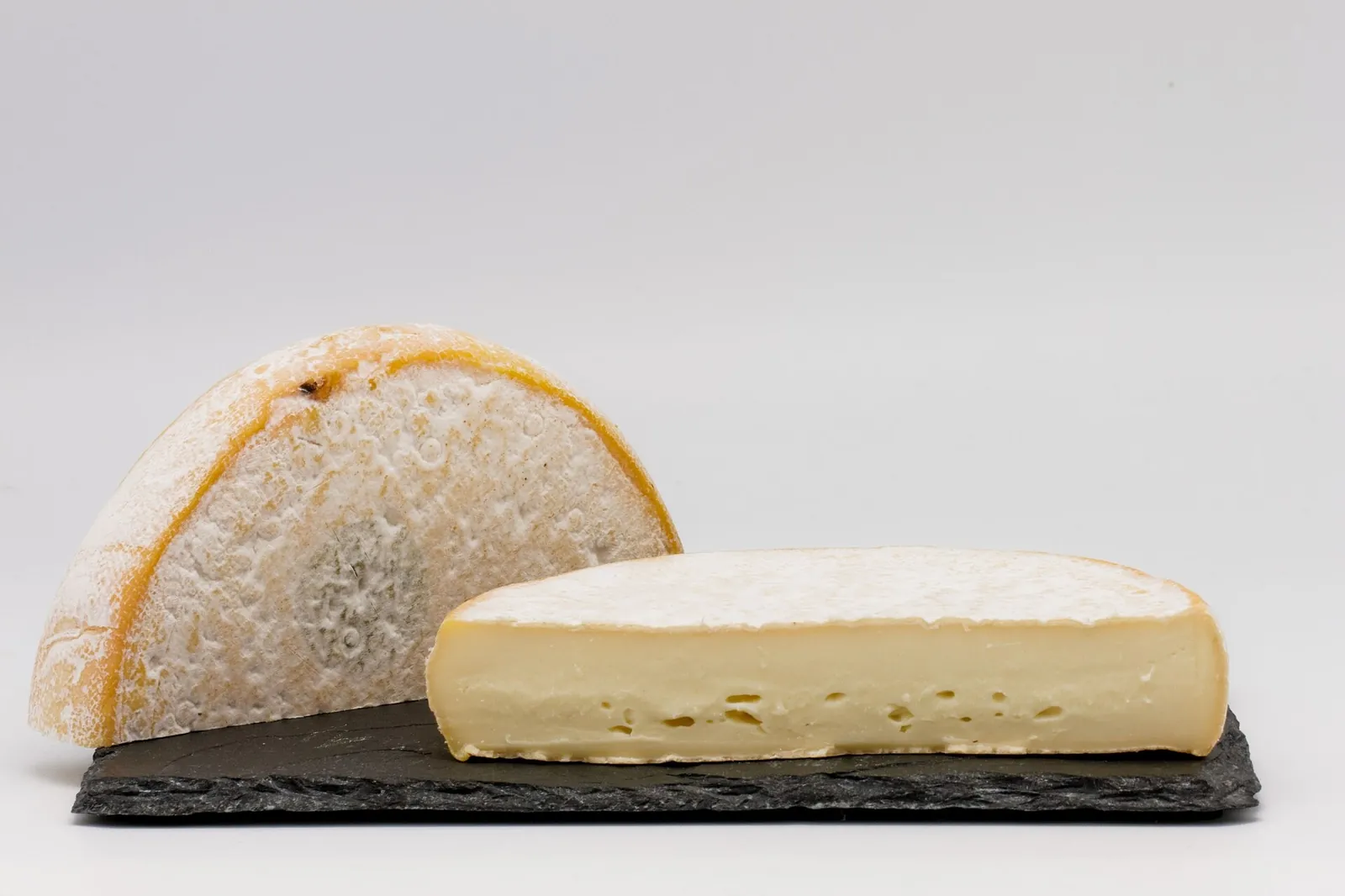 A Reblochon wheel cut open beside a wedge, showing a thin pale orange rind with bloom and a soft ivory paste