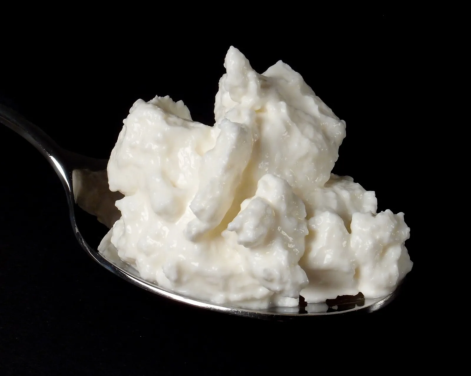 Metal spoon holding a soft, lumpy scoop of white quark against a black background.