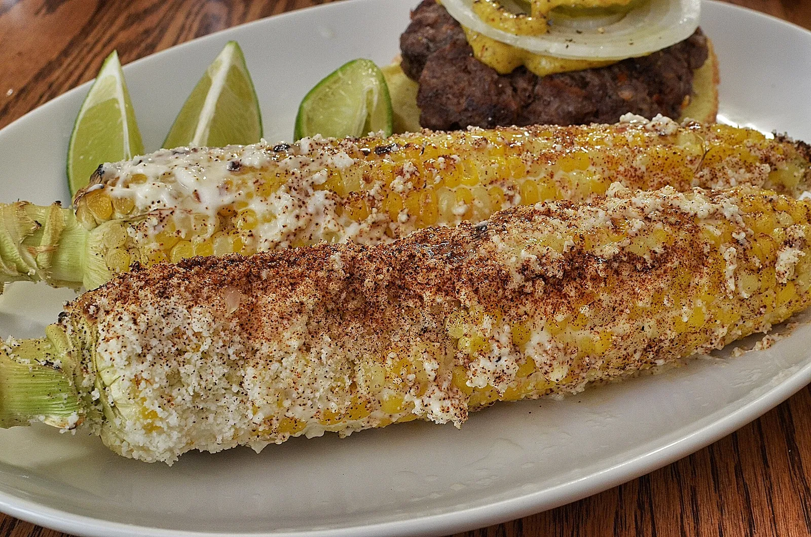 Two grilled ears of Mexican street corn dusted with crumbled cotija cheese and chile, served on an oval plate with lime wedges.