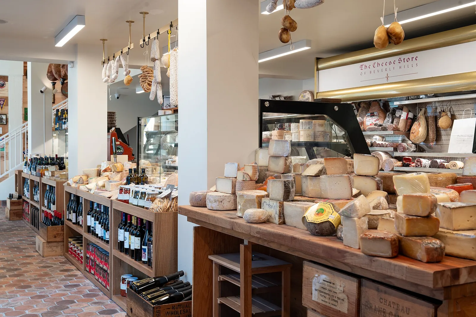 Inside a cheese shop, wheels and wedges of cheese on a wooden counter, with wine shelves and hanging hams behind