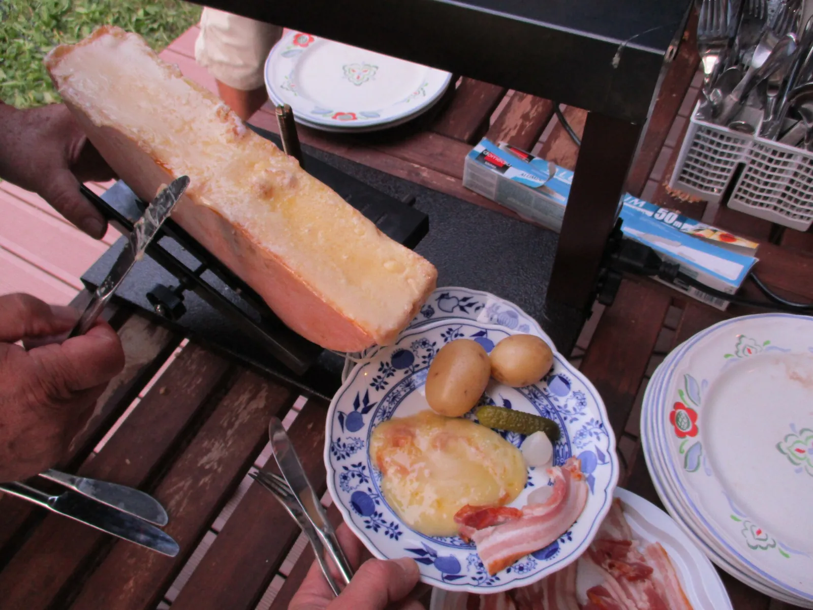 A half wheel of raclette cheese melting under a table heater, with a plate of boiled potatoes, bacon and pickles topped with melted cheese.