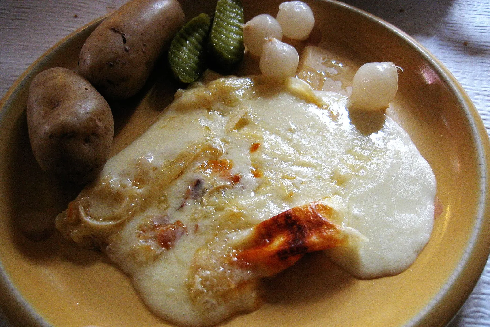 Melted raclette over boiled potatoes on a plate with pickled onions and cornichons