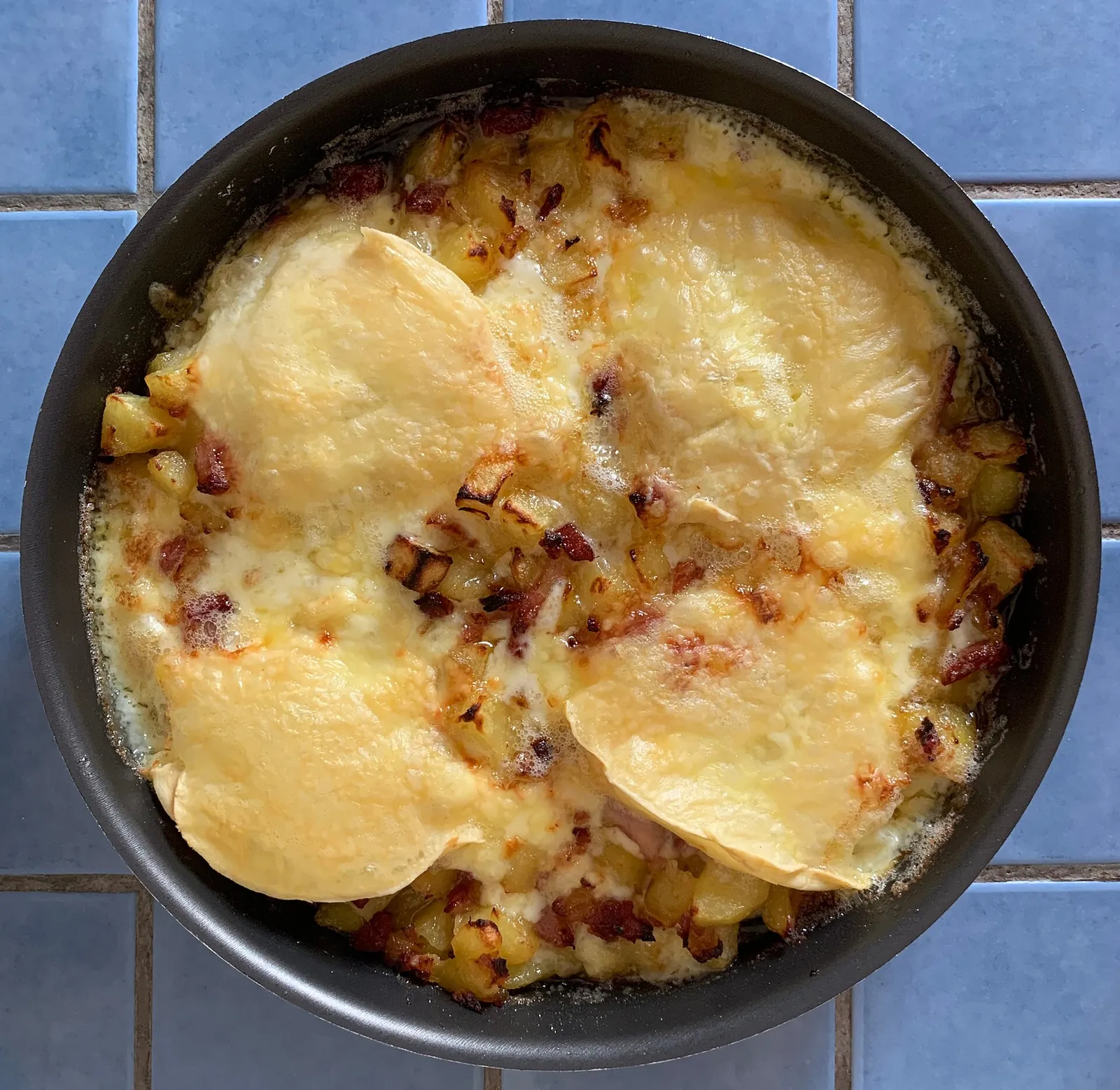 Tartiflette in a black pan with melted Reblochon rounds over potatoes and bacon, set on blue tiles