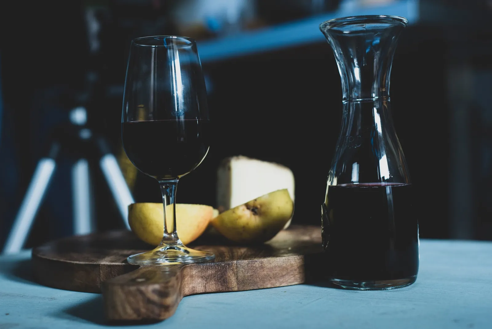A glass and a carafe of red wine on a wooden board with a halved pear and a wedge of firm white cheese behind them.