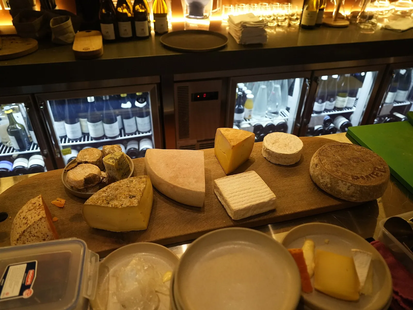 A wooden board of French cheeses, including wheels, wedges and a square washed-rind cheese, on a counter with a wine fridge behind.