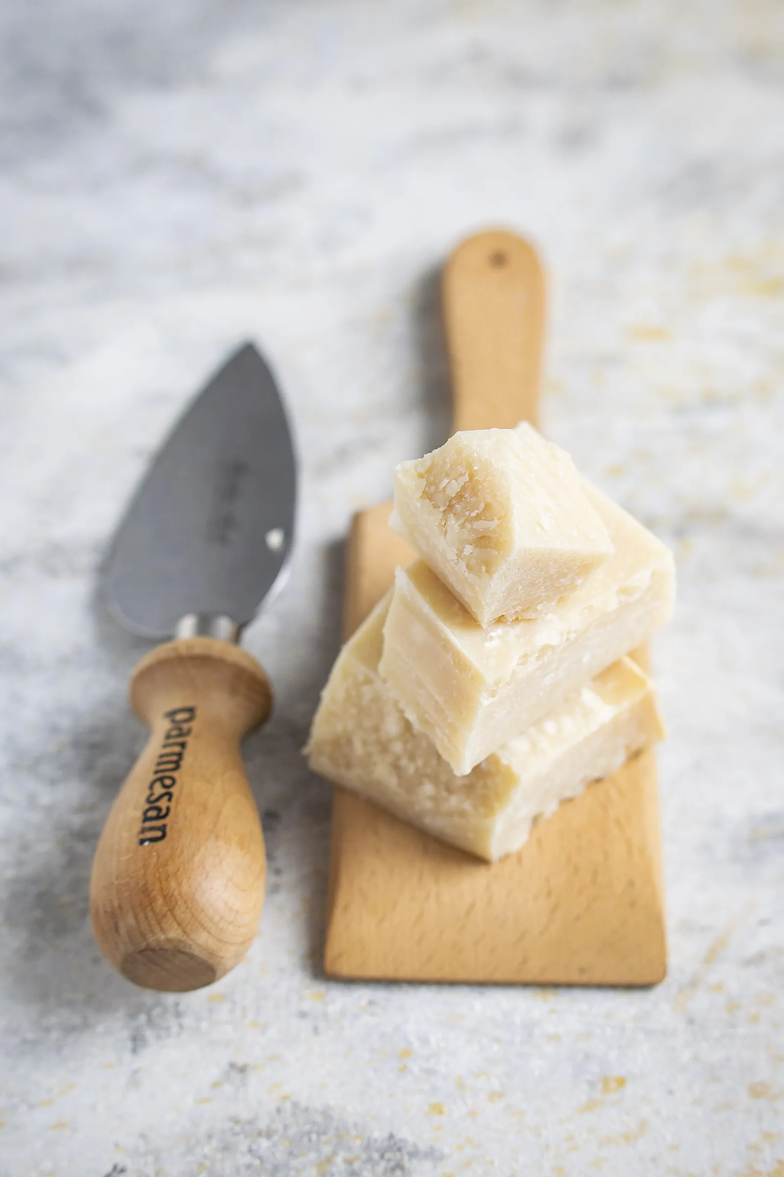 Crumbly pieces of hard aged cheese stacked on a small wooden board beside a pointed cheese knife, on a pale stone surface.