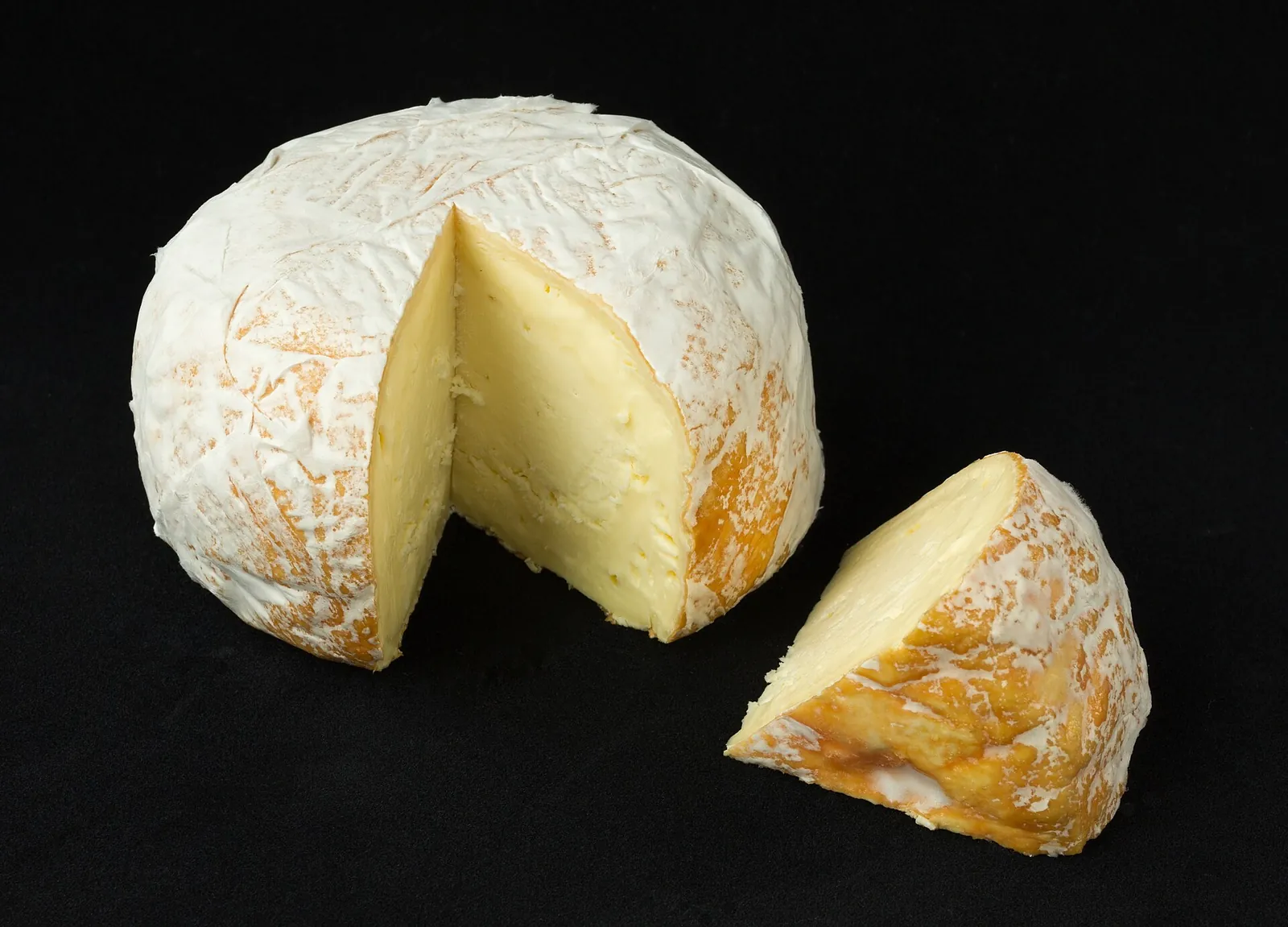 Whole washed rind cheese with a cut wedge on black, showing its pale rind dusted with white mold and orange patches.