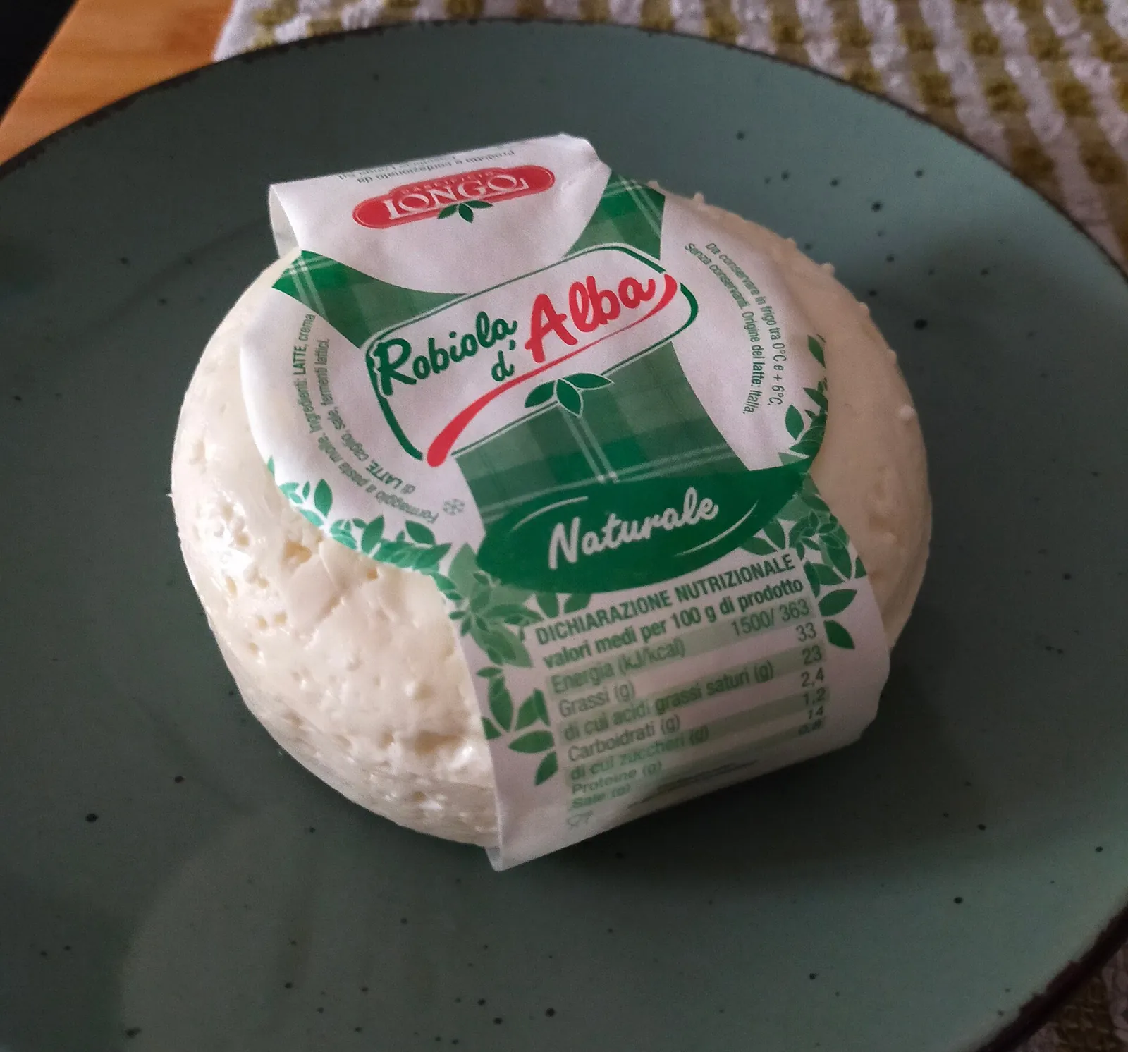 A round Robiola d'Alba cheese partly wrapped in green and white paper on a dark green plate