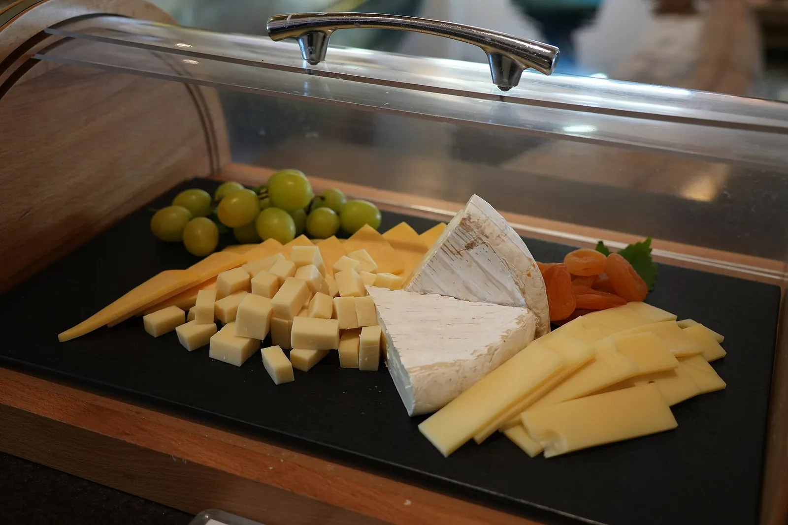 A black slate tray under a clear roll-top lid holding cubed and sliced cheese, a white-rind round, grapes and dried apricots.