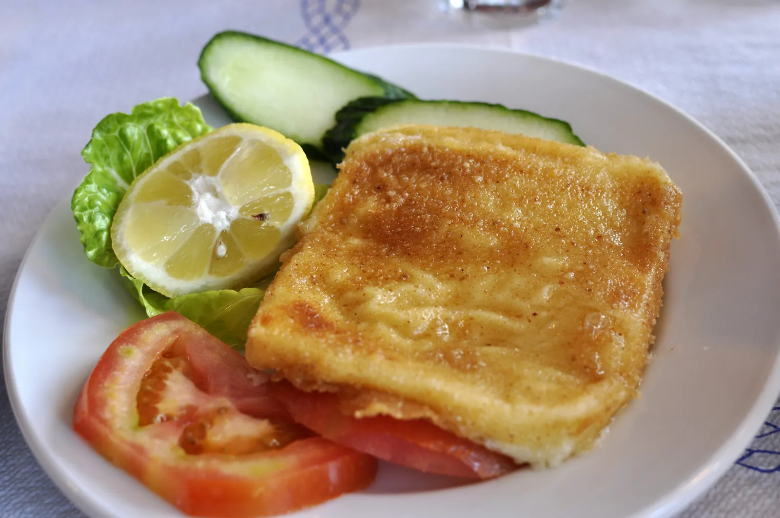 A golden pan-fried slice of Greek saganaki cheese on a white plate with lemon wedge, tomato slices, lettuce and cucumber