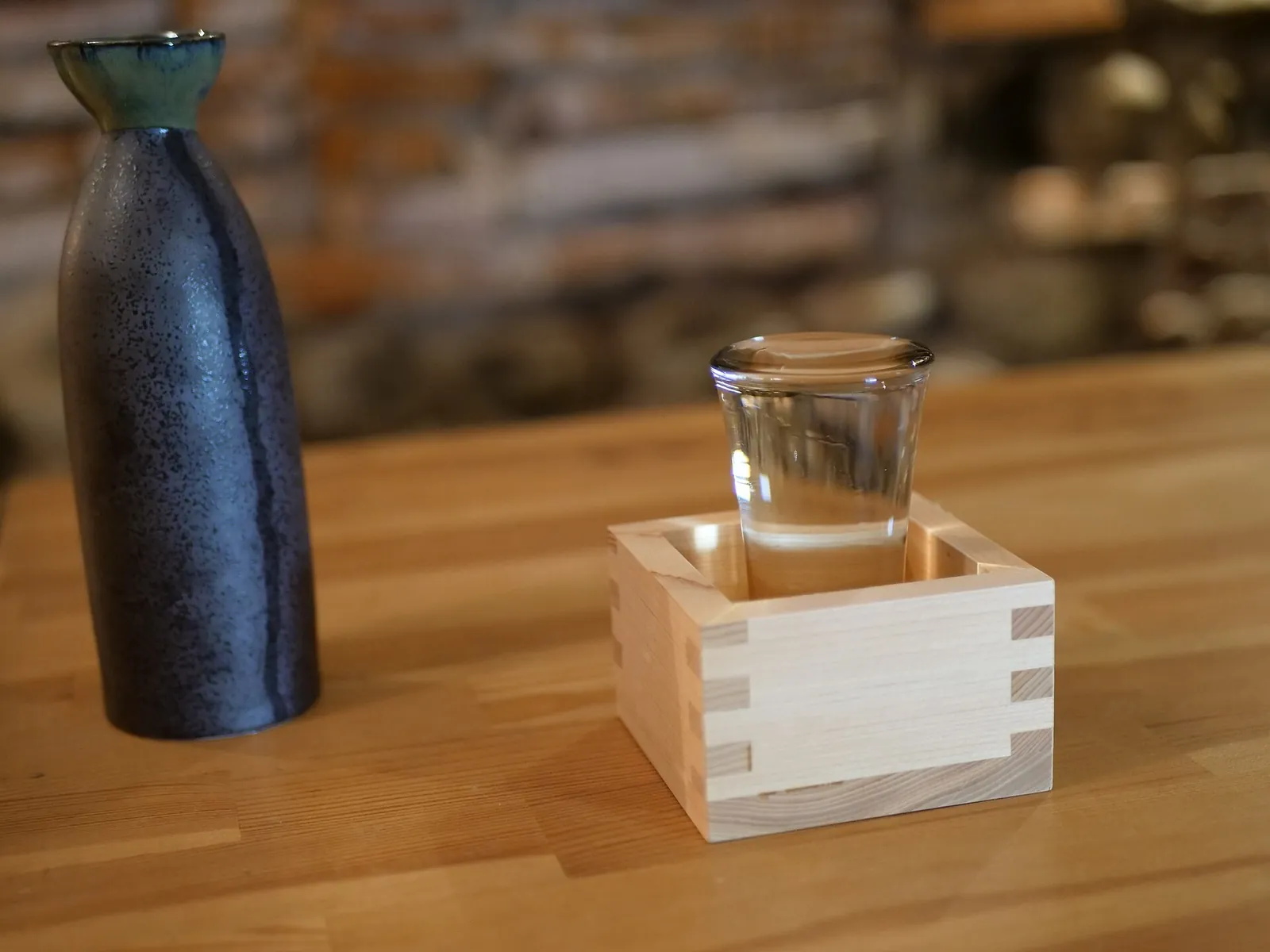 A black ceramic sake flask beside a small glass of sake sitting in a wooden masu box on a wooden counter.