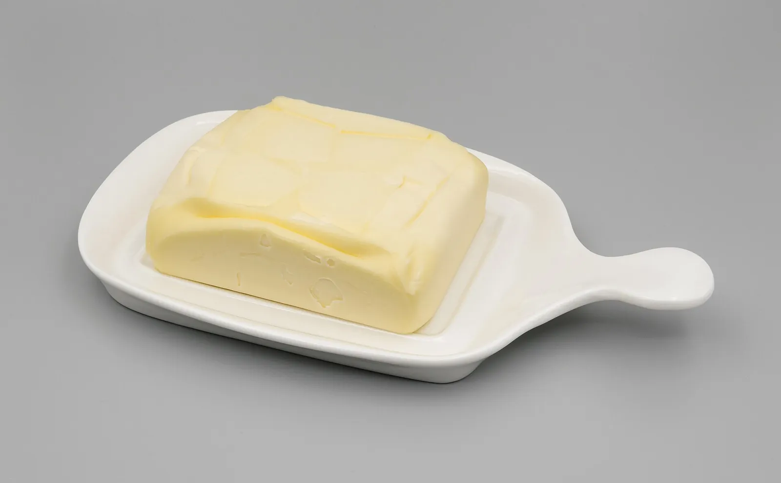 A block of pale yellow butter sitting in a white ceramic butter dish with a handle, on a gray background.