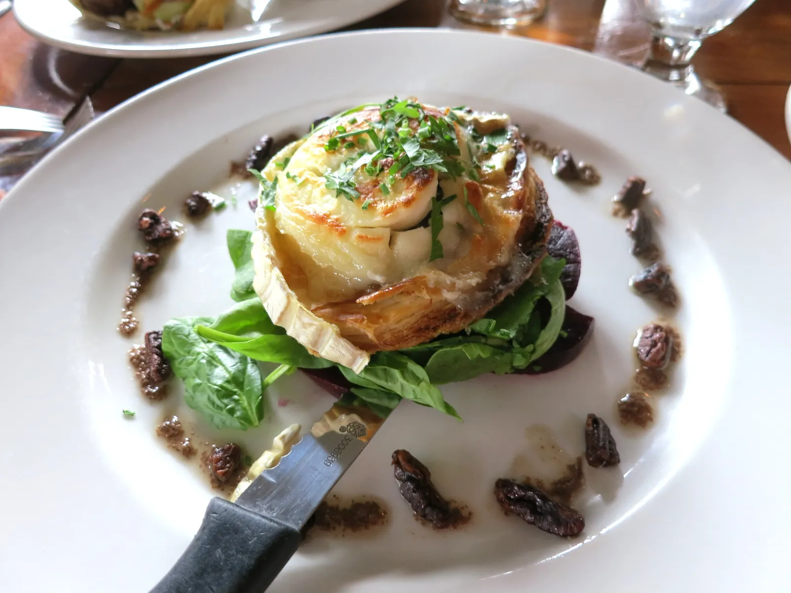 A small baked goat cheese tart topped with fresh herbs, served on a white plate with greens and dark mushroom pieces