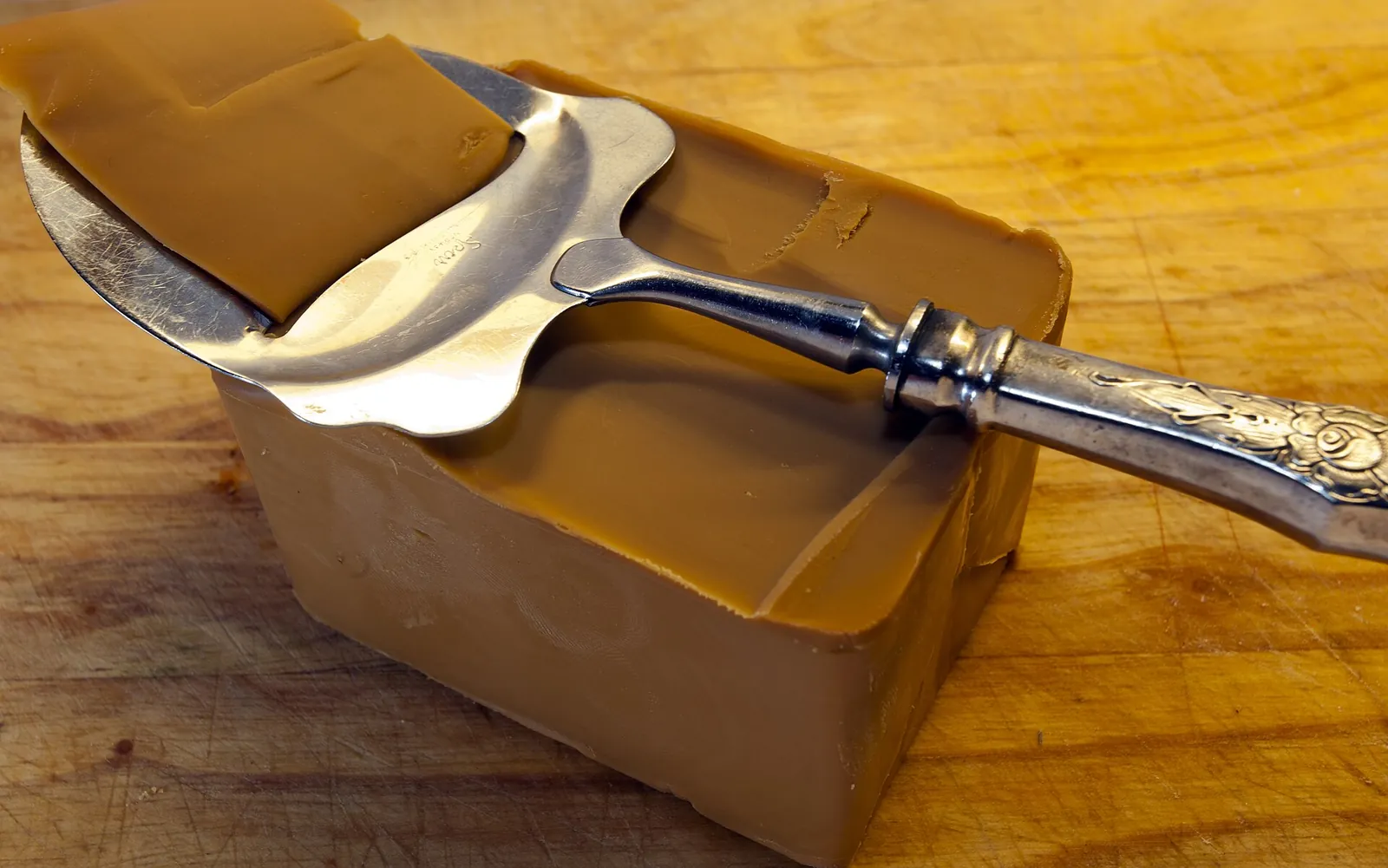 A block of brown Norwegian goat cheese on a wooden board, with a silver cheese slicer shaving a thin slice from its top.
