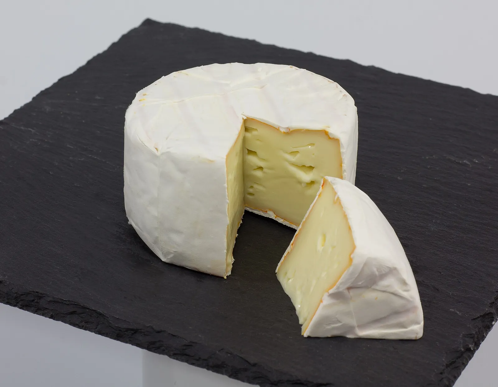 A round Alpine-style cheese with a wedge cut out, showing a pale interior with small holes, on a black slate board