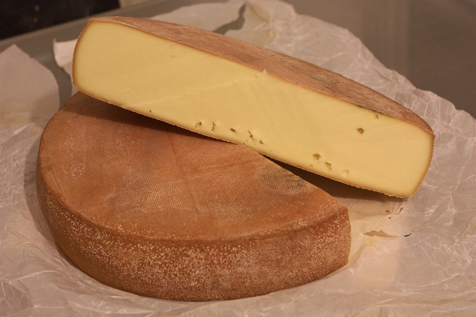 A half wheel of cheese with a golden brown rind, with a long wedge of the same cheese resting on top, on white paper.