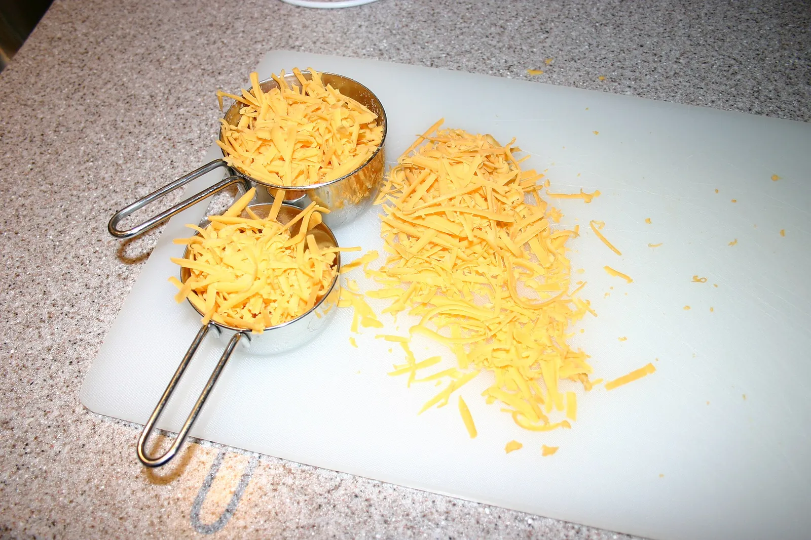 Freshly shredded orange cheddar cheese in two metal measuring cups and spilled across a white cutting board.