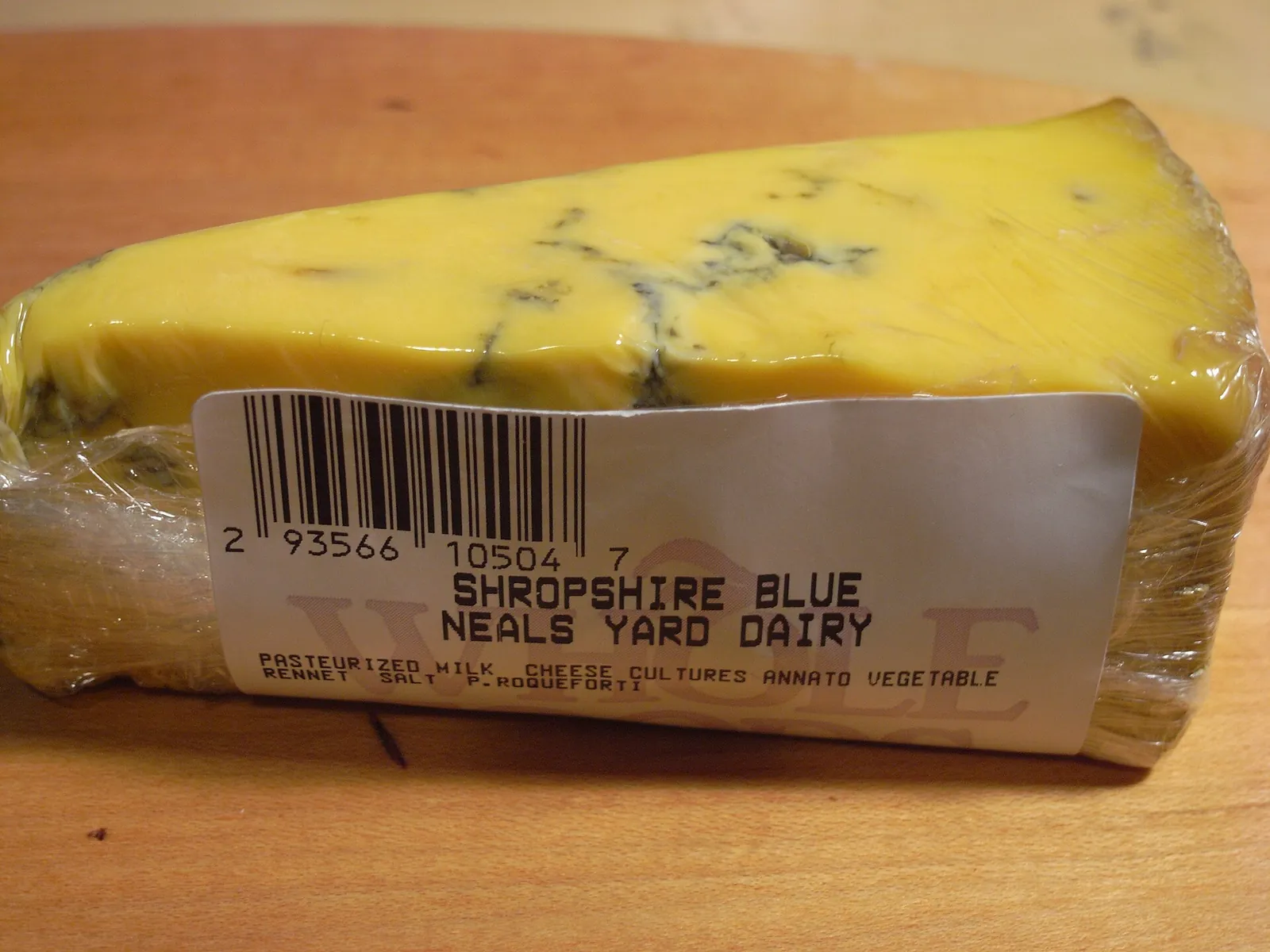 A wedge of Shropshire Blue with orange paste and blue veins, wrapped in plastic on a wooden board