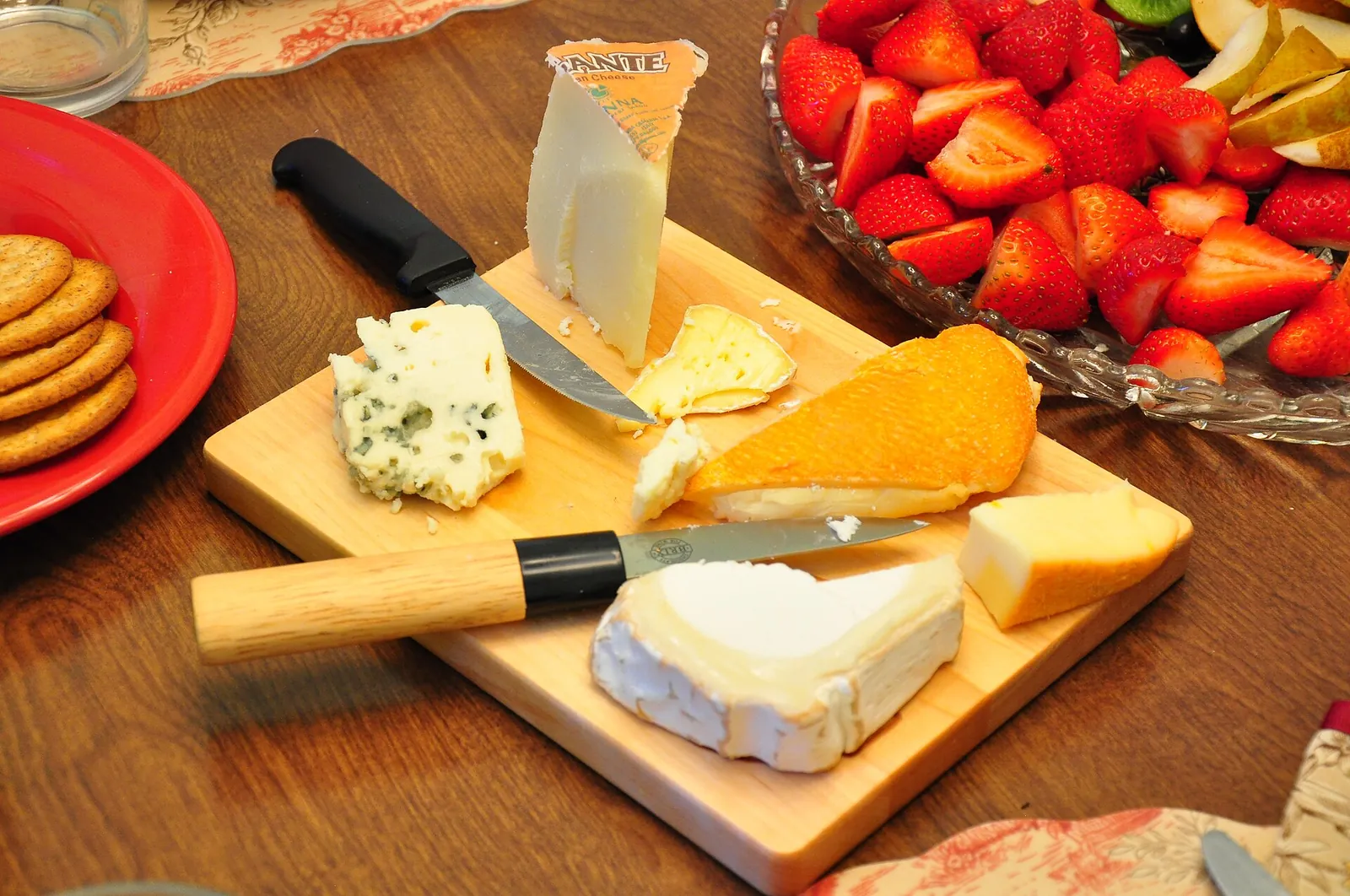 Wooden board of cheeses with different textures, including blue, soft bloomed rind, semi soft orange and firm wedges, beside crackers and strawberries.