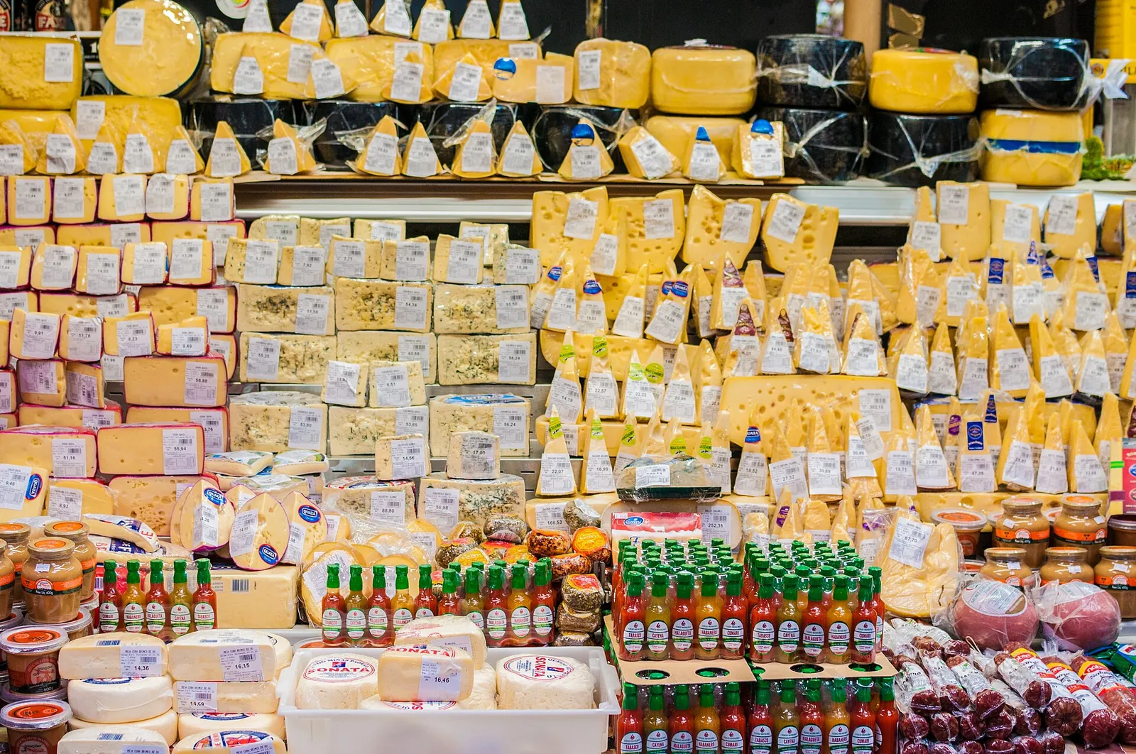 A cheese stall in the Municipal Market of São Paulo, Brazil, with stacked wedges and wheels of cheese, hot sauce bottles and sausages below.