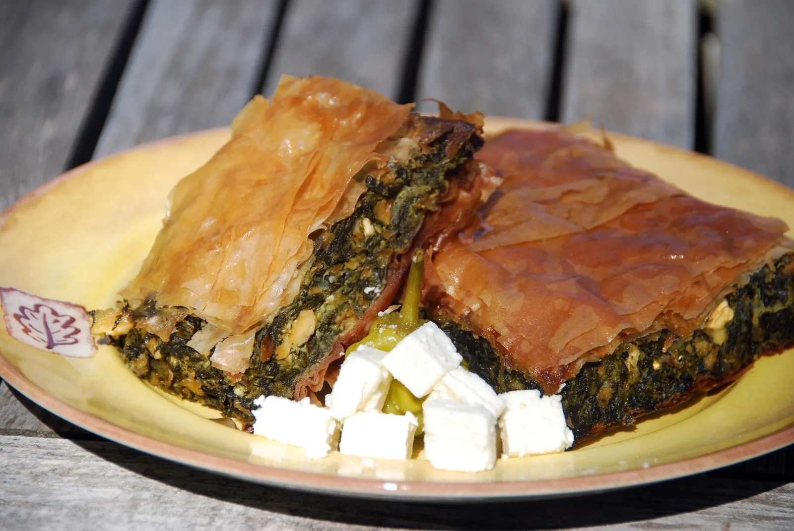Two wedges of spanakopita showing crisp flaky phyllo layers around a green spinach and feta filling, with cubes of feta on a plate