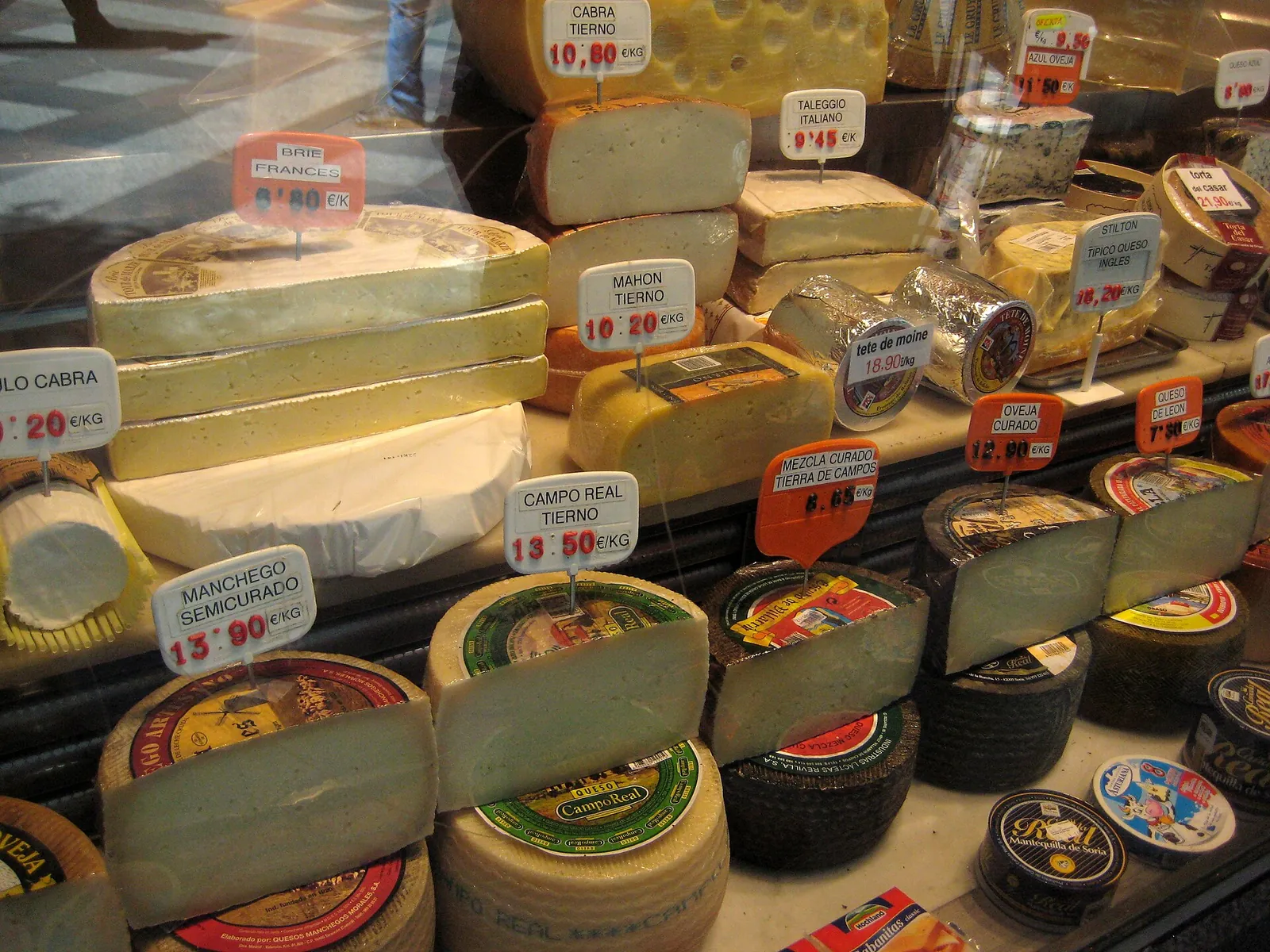 A glass-fronted cheese display at a market in Madrid with Manchego, Mahón and goat cheeses, each marked with a price per kilogram in euros.