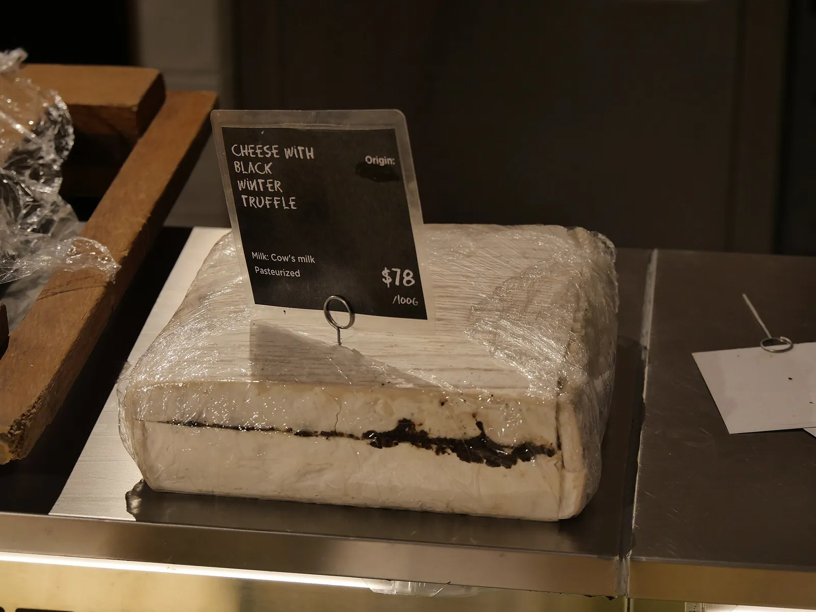 A square wedge of white cheese with a dark band of black winter truffle through the middle, wrapped in plastic beside a price sign