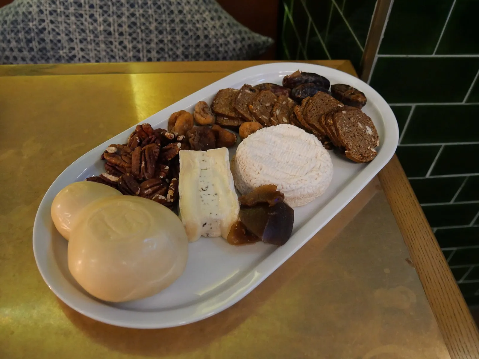 An oval white plate with a round of soft cheese, a wedge of blue cheese, two golden rounds, pecans, crackers and dried fruit