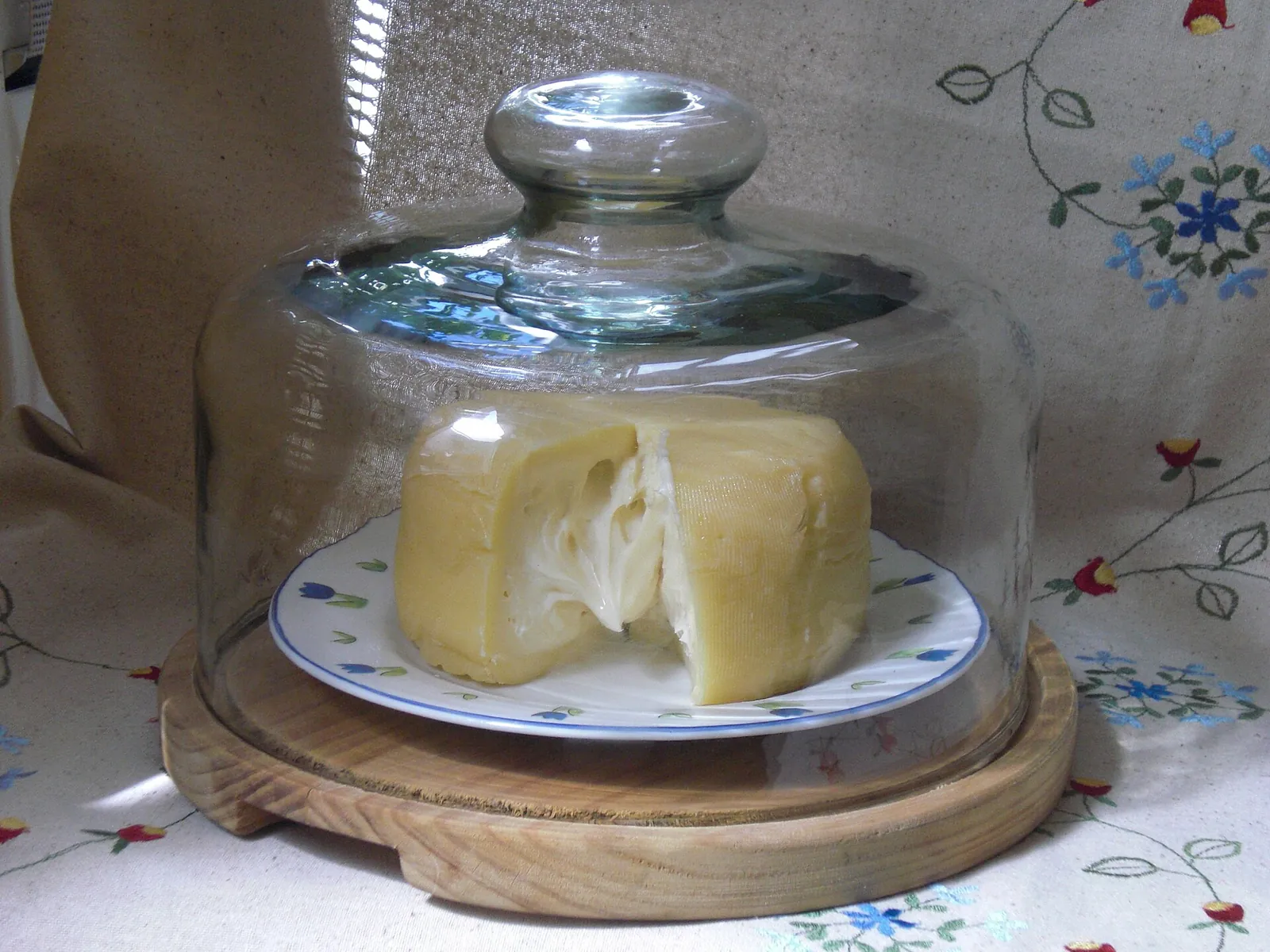 A round cheese with a wedge cut out sitting on a plate under a glass dome on a wooden stand, on an embroidered tablecloth.