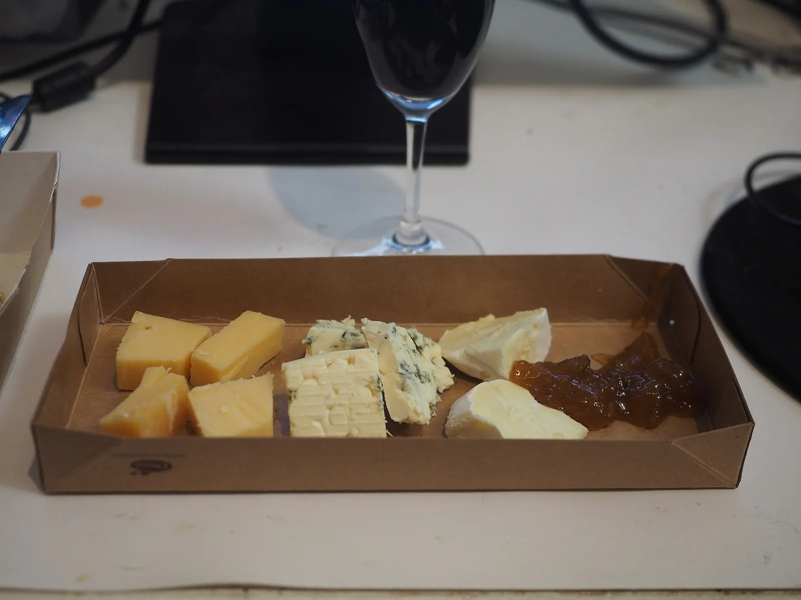 A cardboard box holding cubes of yellow cheese, blue cheese, a soft white cheese and a spoonful of fruit jam, with a wine glass behind