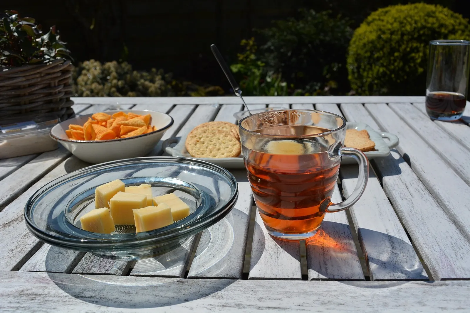 Cubes of yellow cheese on a glass dish beside a glass mug of tea, crackers and a bowl of snacks on a garden table.