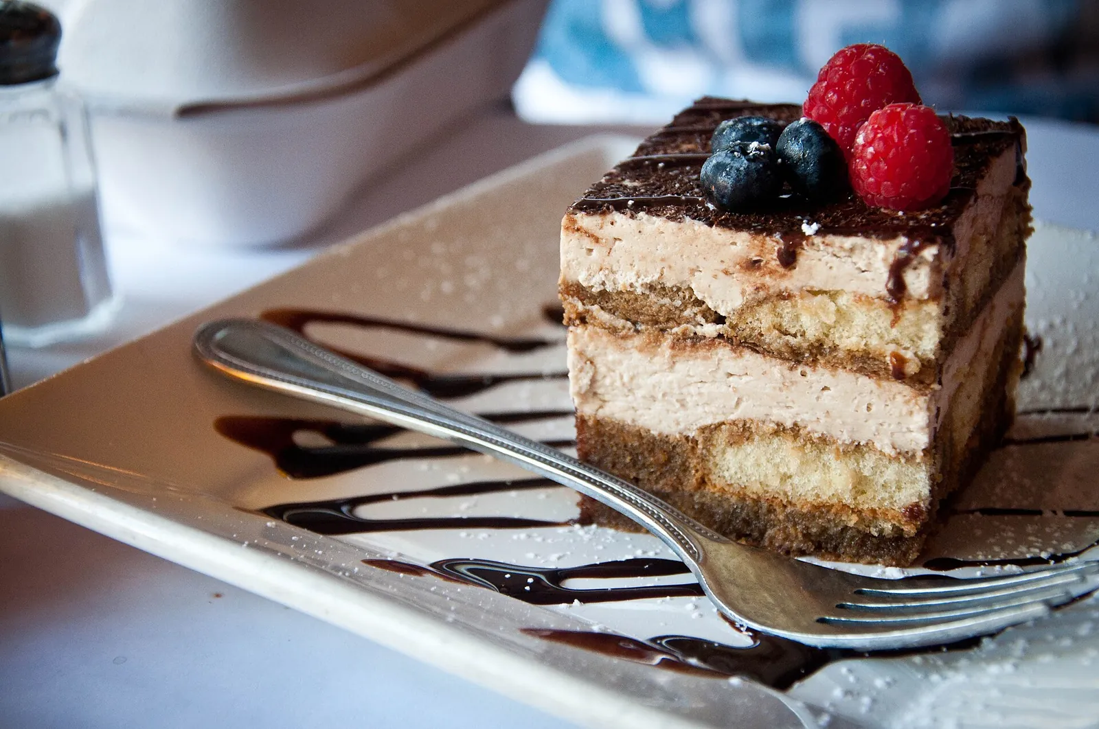 A slice of tiramisu with layers of cocoa-dusted cream and soaked ladyfingers, topped with blueberries and raspberries, on a white plate with chocolate drizzle